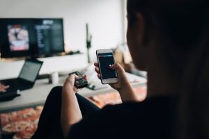 Rear view of teenage girl using phone app and remote control while watching smart TV at home